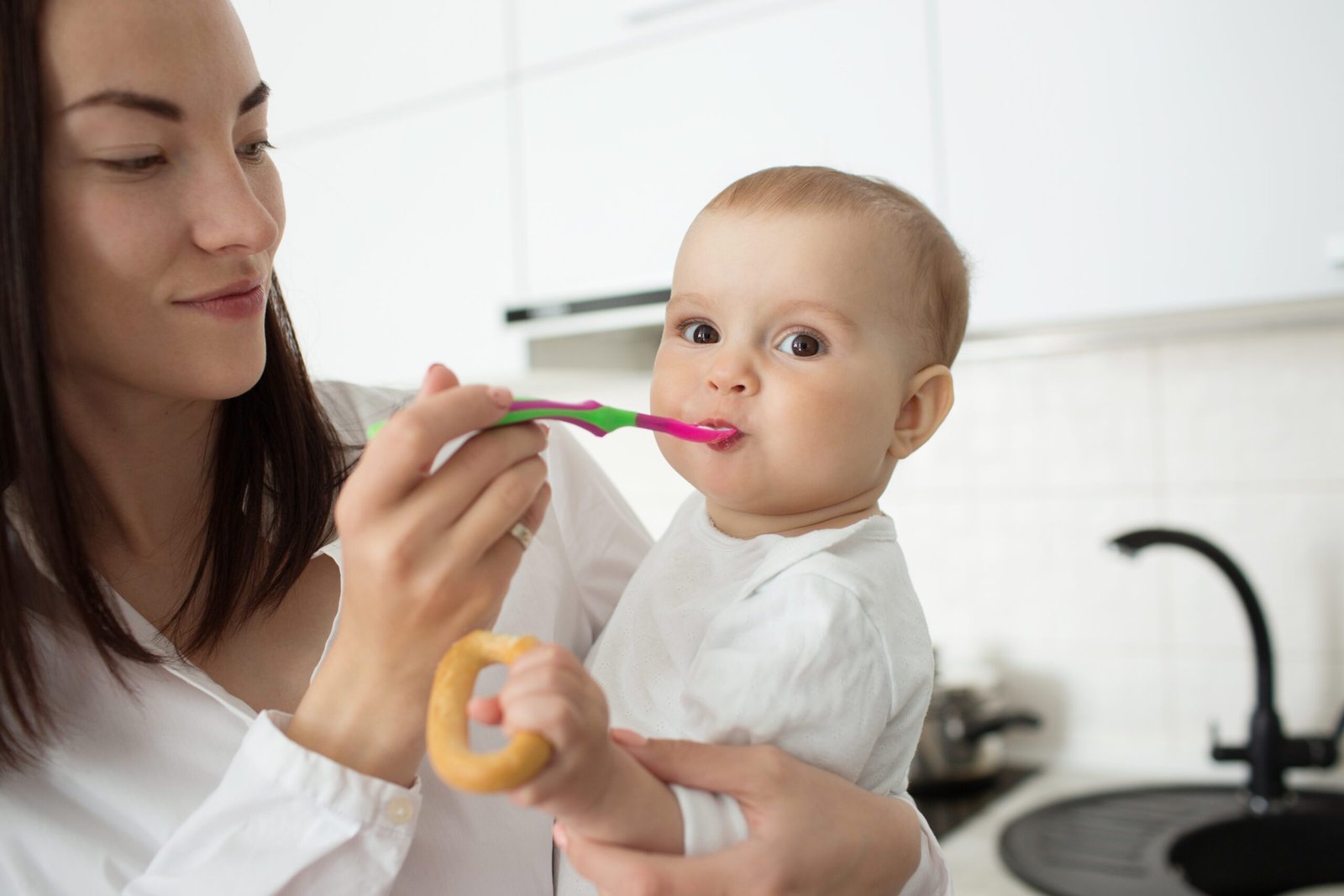 Close up of young handsome mother holding her little son on hands and feeding him, with spoon. Child eating baby food and holding a cookie in hand