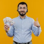 young bearded man in blue shirt with headphones with microphone holding cash showing thumbs up smiling with happy face standing over orange background